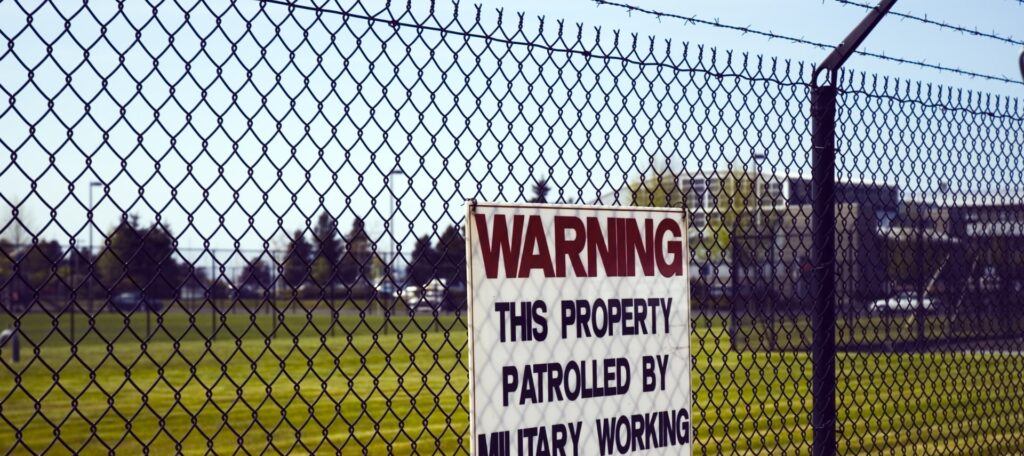 Military base perimeter fence with warning sign and barbed wire security.